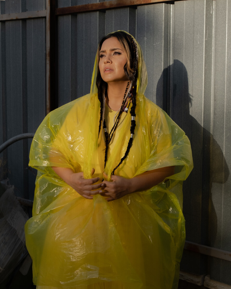 Neta Elkayam porte une robe jaune sous une combinaison en plastique, ses cheveux sont tressés et elle regarde au loin.