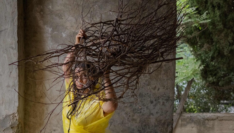 Neta Elkayam porte une robe jaune à moitié en plastique et porte un tas de bâtons de bois sur la tête.
