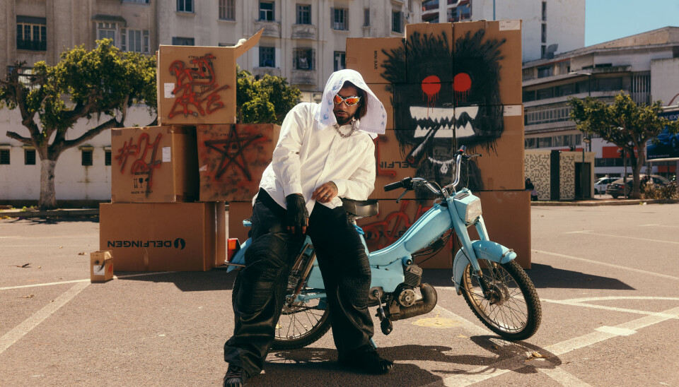 Une personne vêtue d'un sweat à capuche blanc se tient à côté d'un scooter personnalisé bleu clair dans un parking avec des boîtes et des graffitis.
