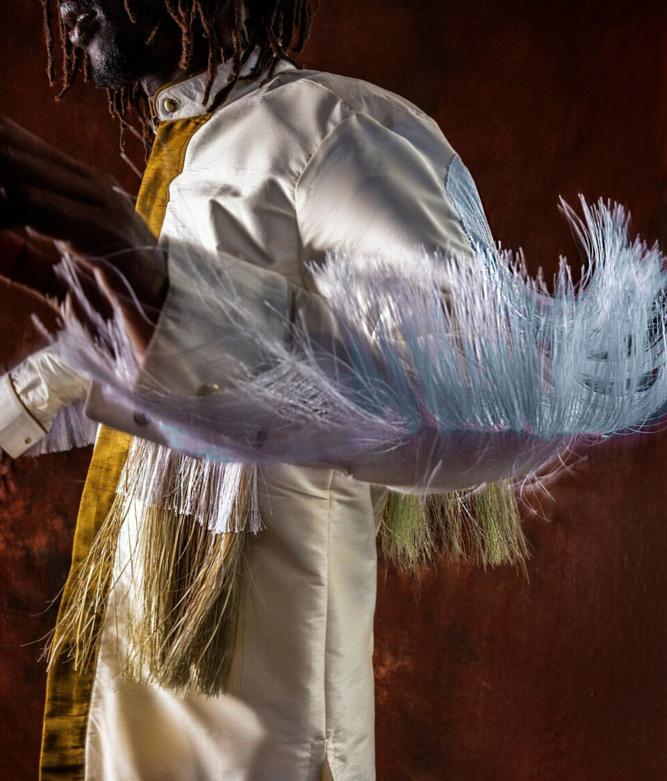 Emmanuel Jal porte une tenue de cérémonie blanche avec des ornements de plumes sur fond sombre.