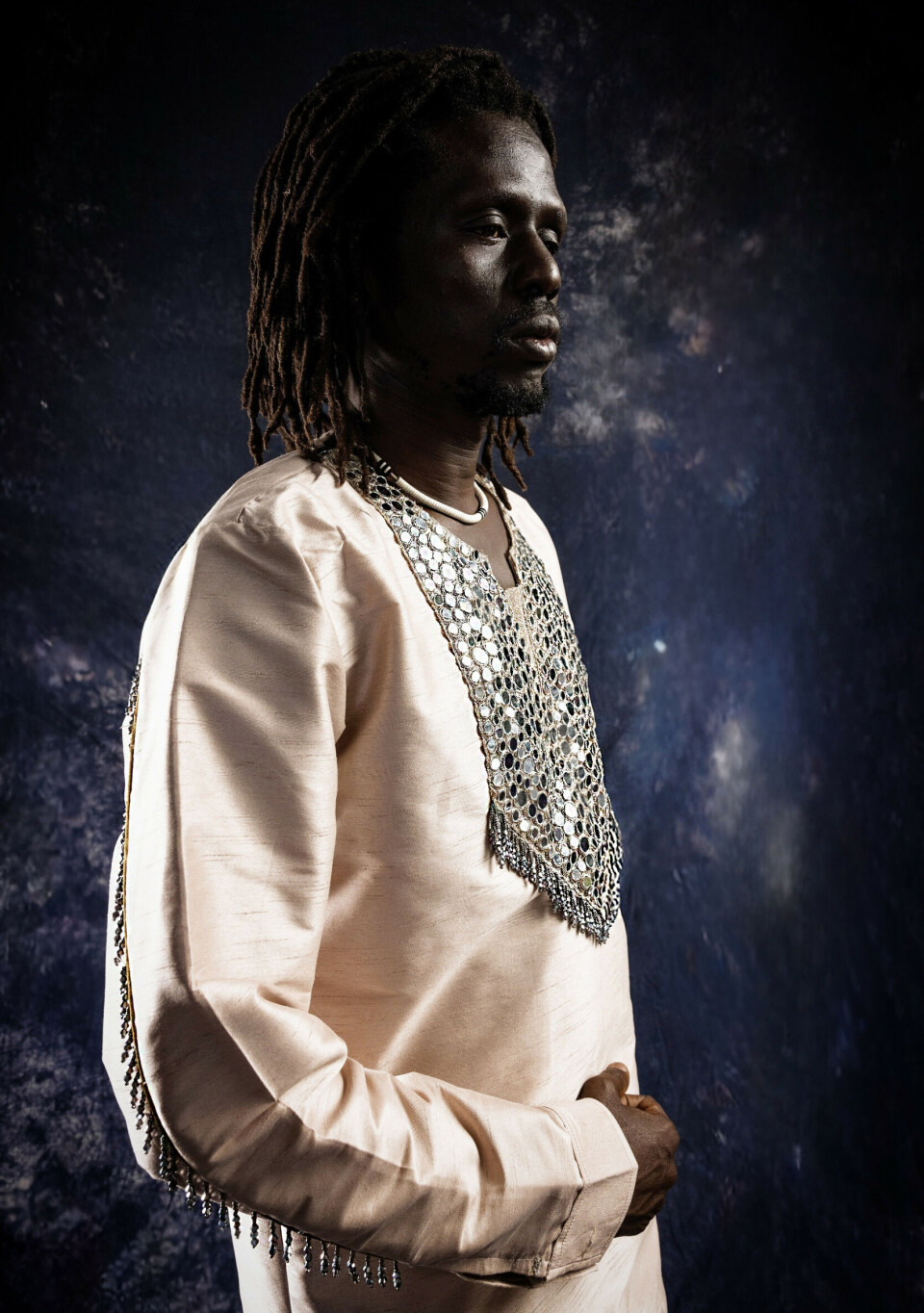 Emmanuel Jal, en tunique brodée crème, pose de profil sur fond sombre.