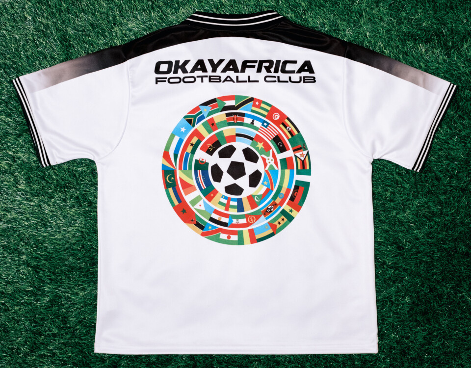 Maillot blanc du Okaya Africa Football Club avec un motif de football multicolore sur herbe verte.