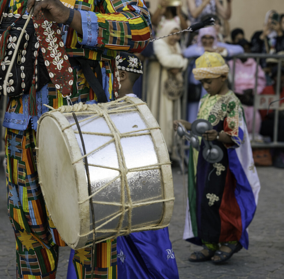 Un homme porte un gros tambour ; derrière lui, un petit garçon tient un grand qraqeb. Tous deux sont vêtus de vêtements traditionnels colorés.