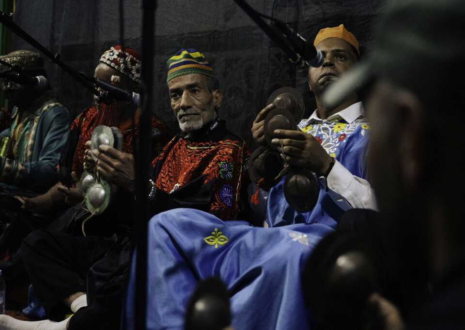 Cinq hommes sont assis par terre devant des microphones, jouant d’instruments et portant des vêtements traditionnels marocains colorés.
