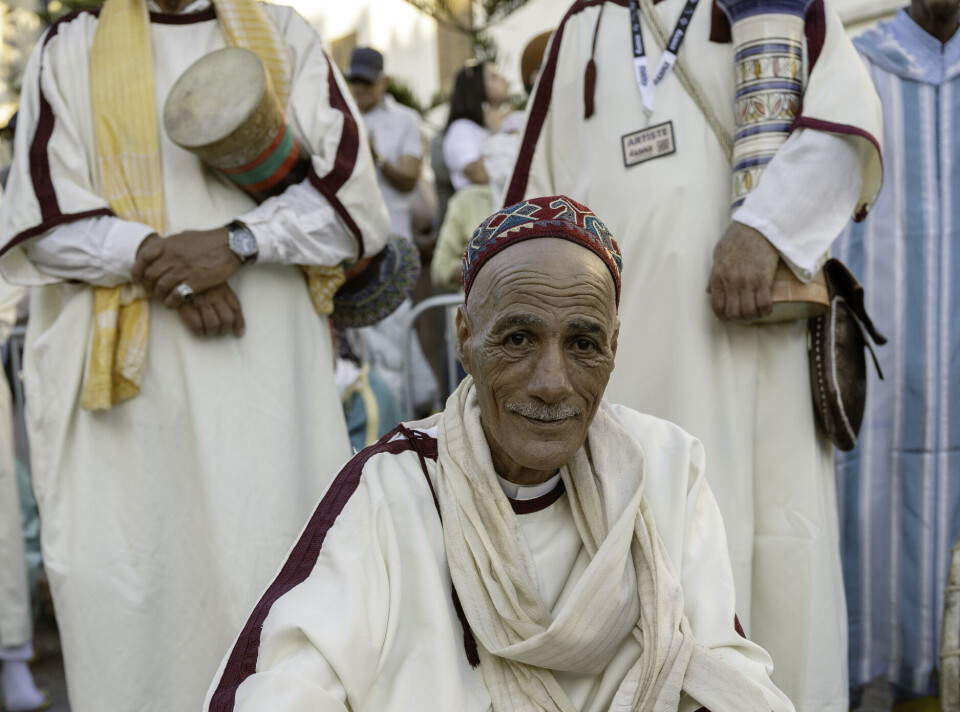 Un homme âgé vêtu d’une tenue marocaine traditionnelle de couleur crème est confortablement accroupi dans la rue et regarde directement la caméra.
