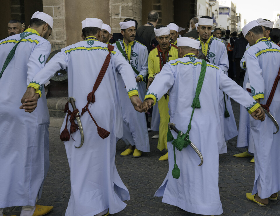 Neuf hommes portant une tenue marocaine traditionnelle bleu clair et des poignards à la taille forment un cercle, se tenant la main et dansant.