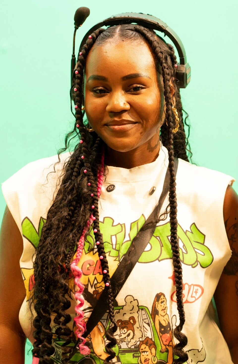 Portrait d'une personne aux longs cheveux tressés portant des écouteurs et une chemise graphique sur fond vert menthe.
