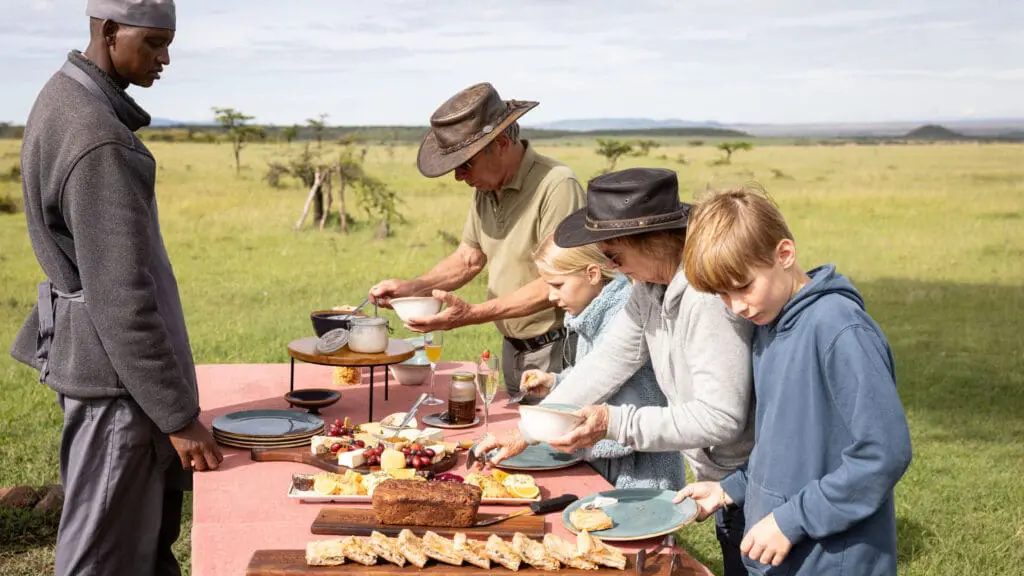 Invités et enfants se servant de la nourriture à une table de petit-déjeuner de brousse située dans une prairie ouverte