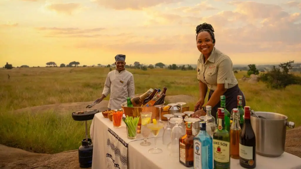 Deux employés du Sayari Camp préparent un safari classique au coucher du soleil sur les plaines ouvertes, avec une table de boissons, de verres et de collations au coucher du soleil, des prairies dorées s'étendant au loin sous un ciel chaud du soir.