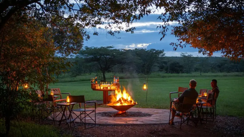 Les invités du camp Encounter Mara sont assis autour d'un feu de camp ouvert au crépuscule, dégustant des boissons et la lumière d'une lanterne surplombant la savane ouverte et les arbres.