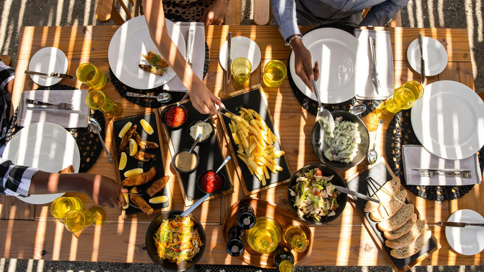 Vue aérienne des invités partageant un repas décontracté en plein air, avec des assiettes, des salades, du pain et des boissons disposés sur une table en bois sous un soleil tacheté.