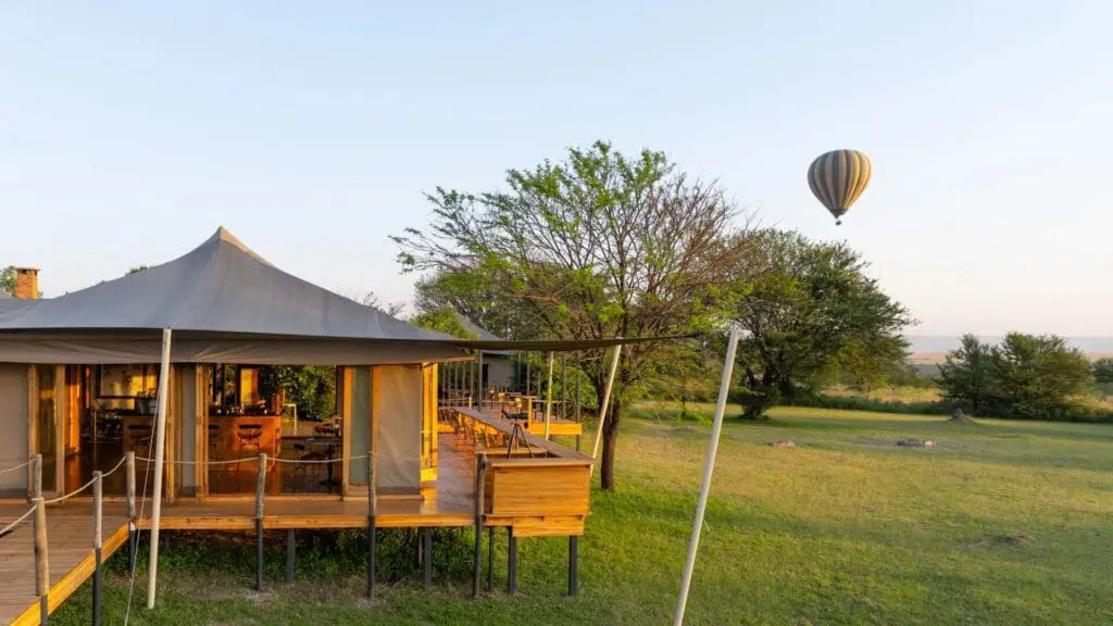 Le salon surélevé et la terrasse du Sayari Camp surplombent les prairies ouvertes du Serengeti, avec une montgolfière flottant au loin aux premières lueurs du matin, encadrées par des arbres et un ciel dégagé.
