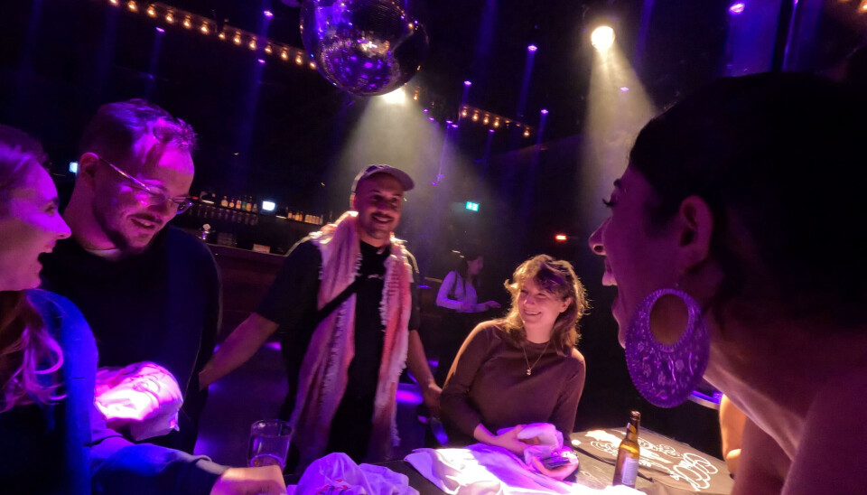 Une femme se tient près d’une table et rit avec un groupe de personnes en face d’elle.