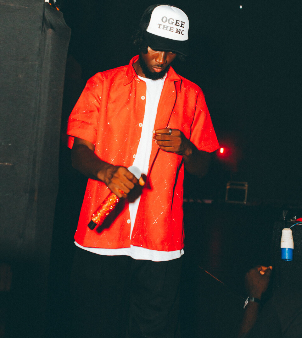 Un artiste en chemise rouge et casquette blanche tient un microphone sur une scène sombre.